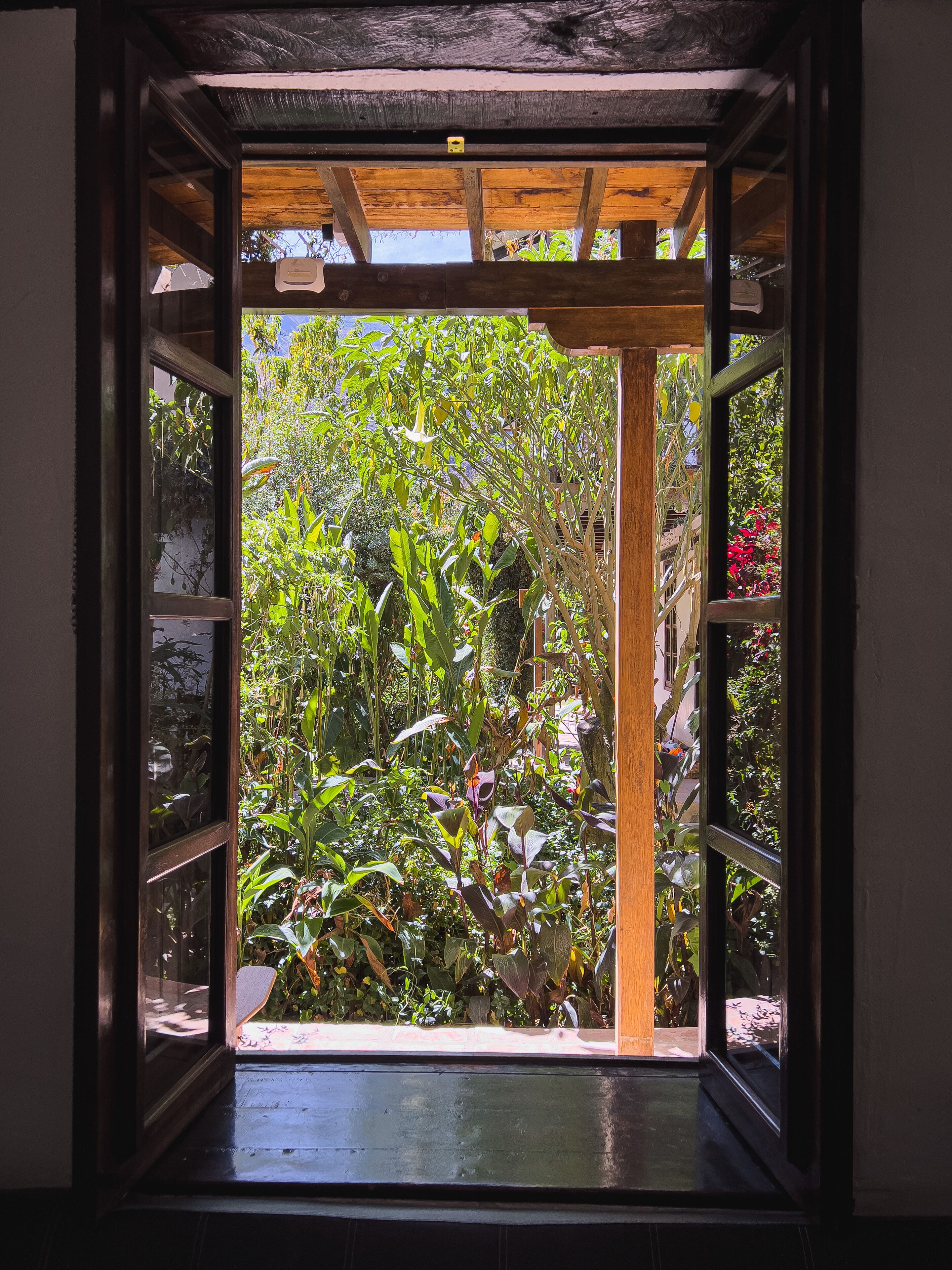 An open wooden door leading to a lush garden with various plants and a wooden structure above.