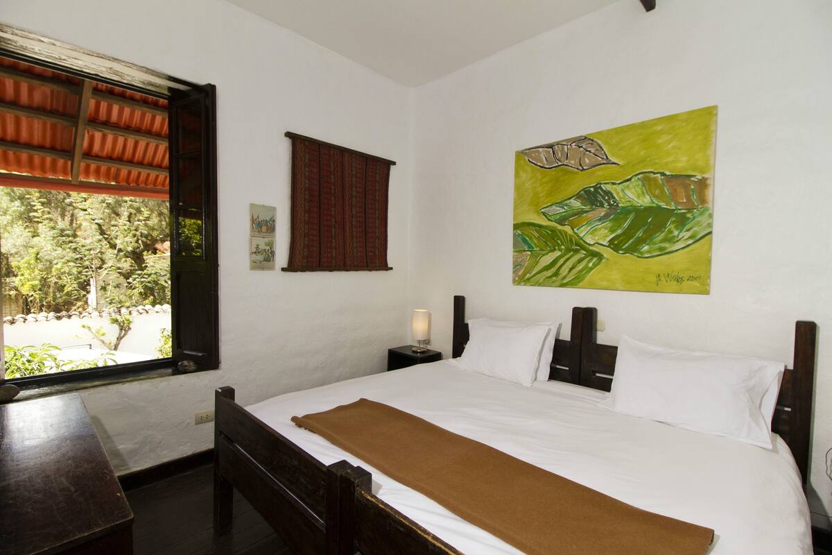 Een slaapkamer met een bed, een raam, een nachtkastje en een schilderij aan de muur.