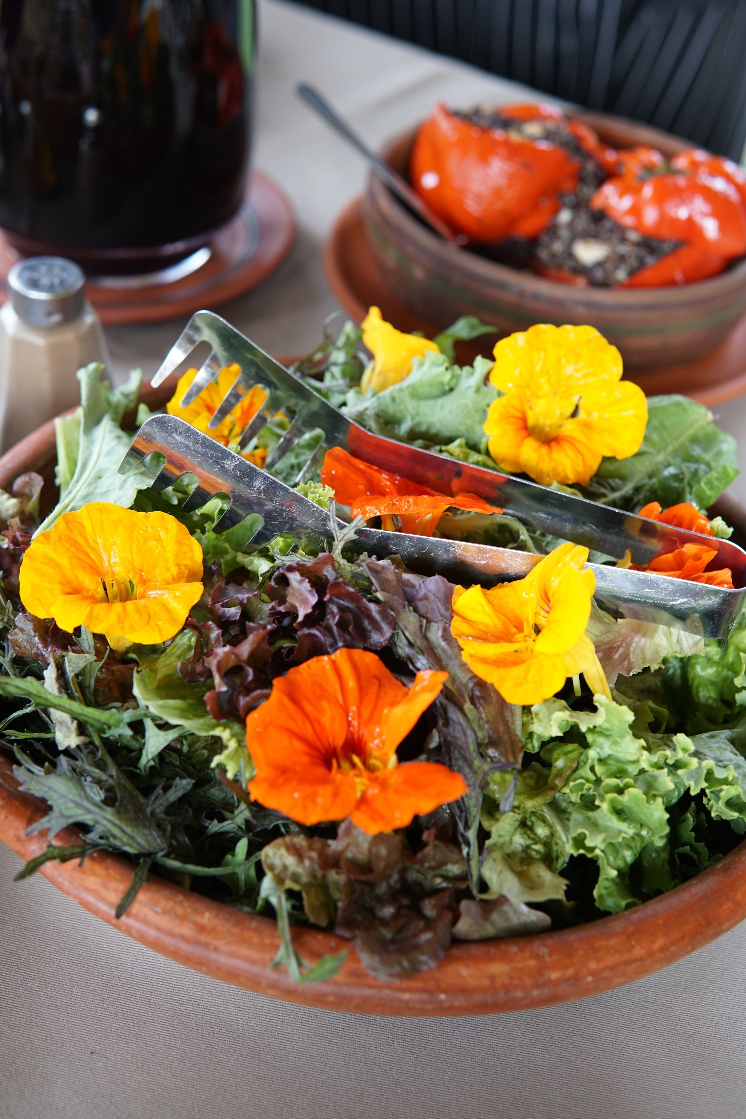 A salad with mixed greens and orange flowers, a fork, and a bowl of tomatoes in the background.