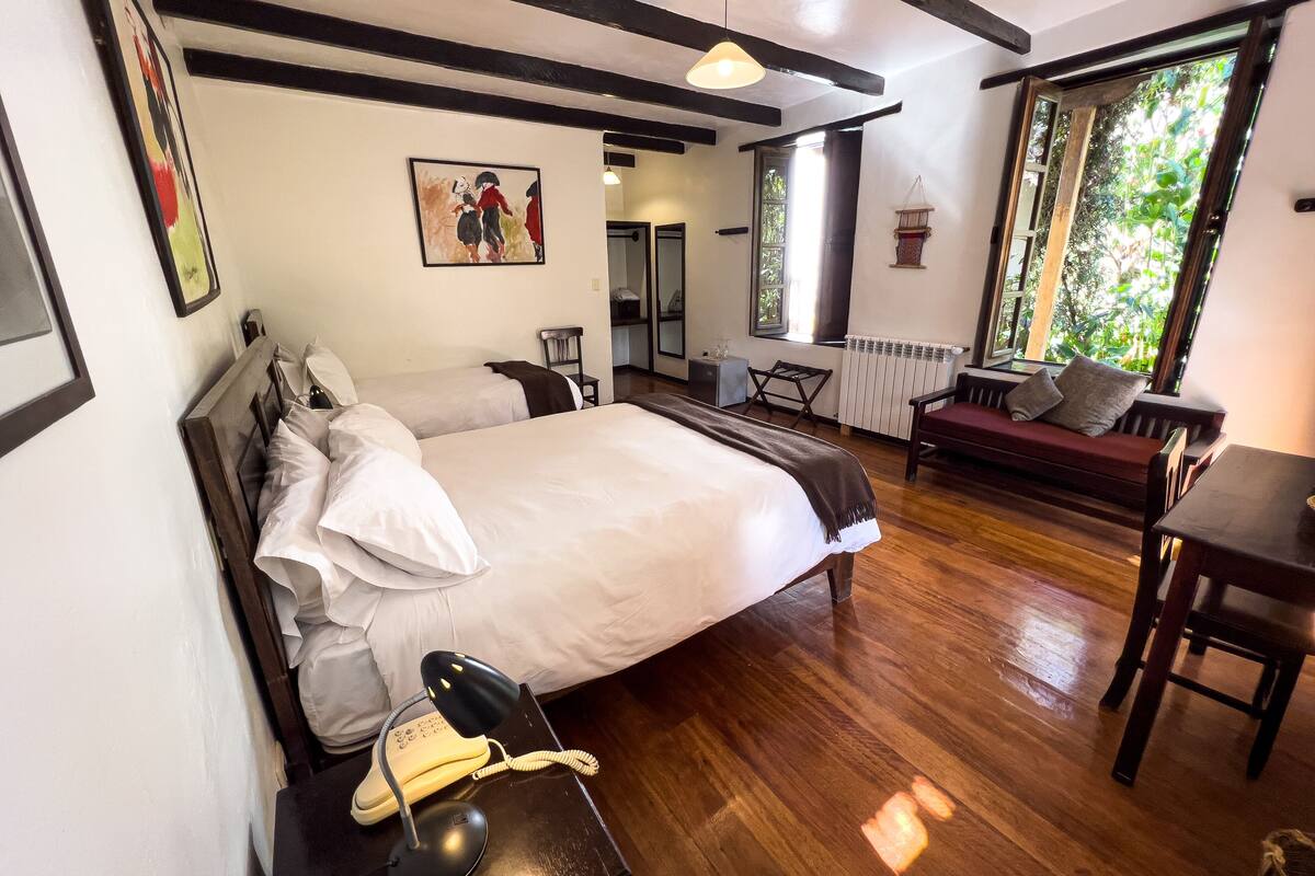 Een slaapkamer met een groot bed, nachtkastjes, een bureau, een stoel, een bank en een raam met uitzicht op groen.