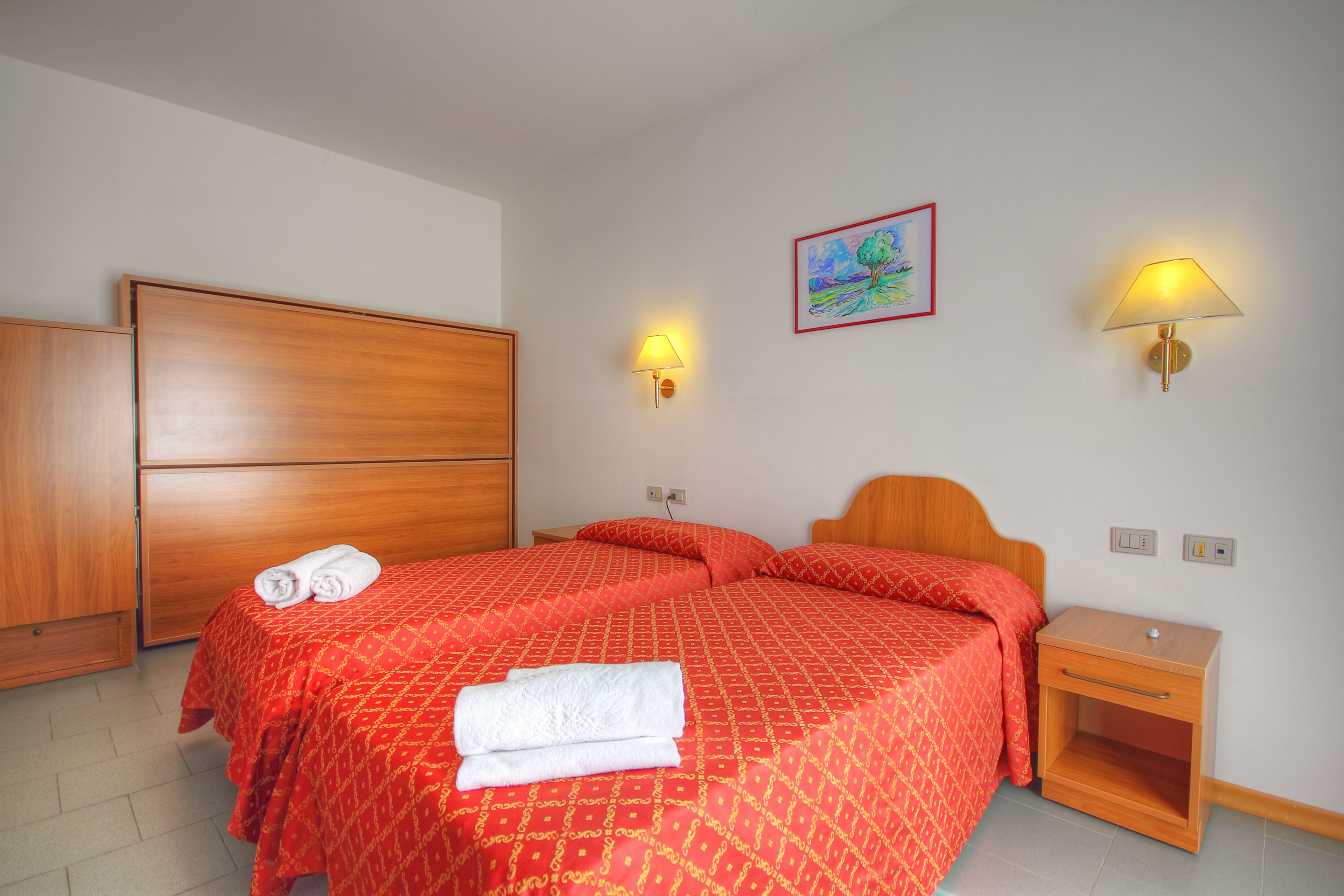 A double bed with red bedding, two bedside tables, a wooden wardrobe, and a painting on the wall.