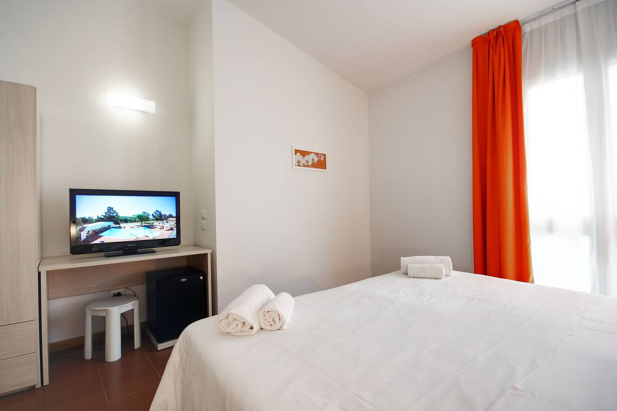 A hotel room with a bed, a TV on a stand, a small stool, and a white towel on the bed.