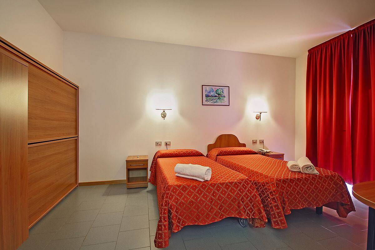A hotel room with two beds, a wooden wardrobe, a small bedside table, and a red curtain.