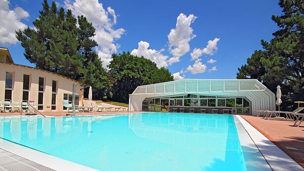A swimming pool with a clear blue water surface, surrounded by lounge chairs and a covered area with a glass roof. - I Girasoli (Lucignano)