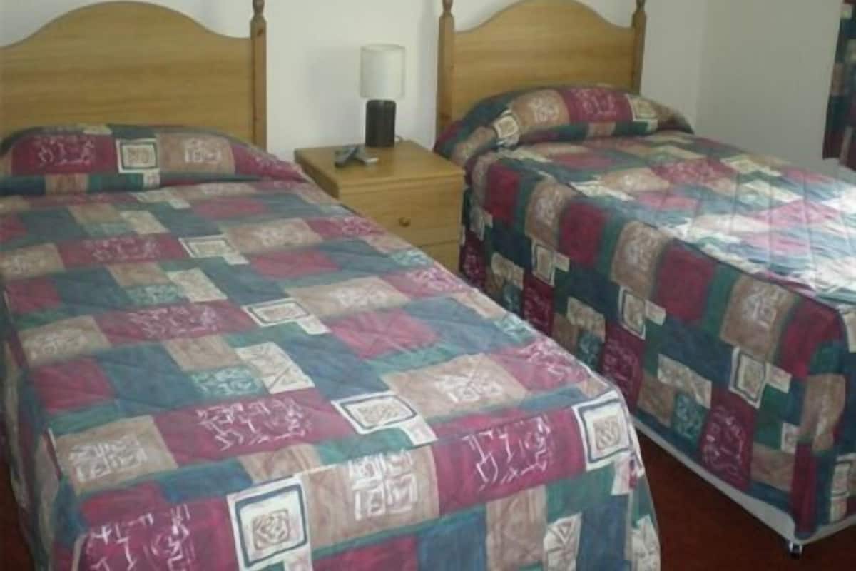 Quarto com duas camas de madeira, cada uma com colcha acolchoada. Há uma mesinha de cabeceira com abajur entre as camas.