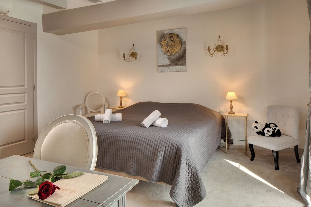 Een hotelkamer met een groot bed, twee bedlampen, een stoel, een tafel met een roos en een kunstwerk aan de muur.