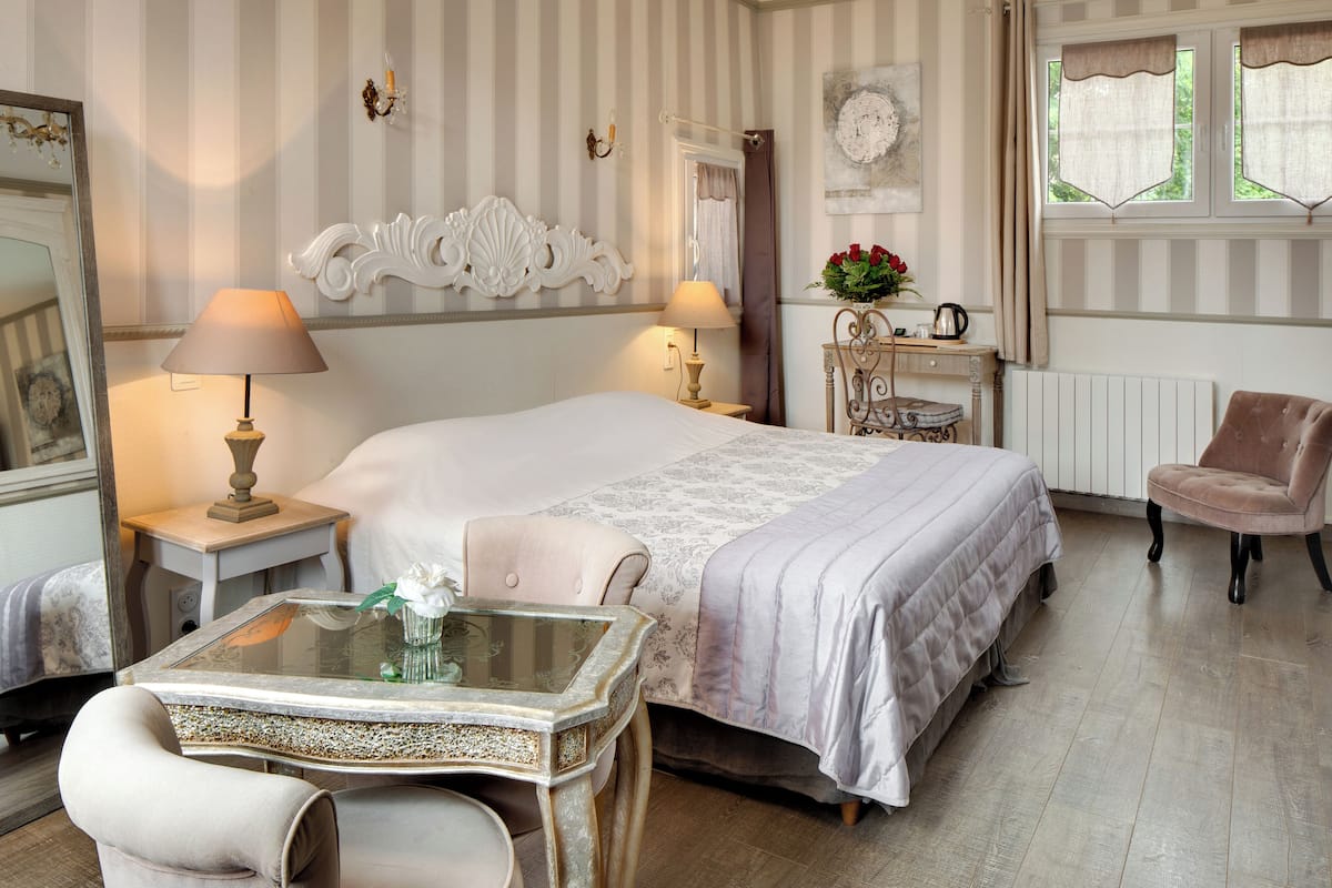 Een hotelkamer met een groot bed, twee nachtkastjes met lampen, een tafeltje met een vaas, een stoel en een raam met gordijnen.