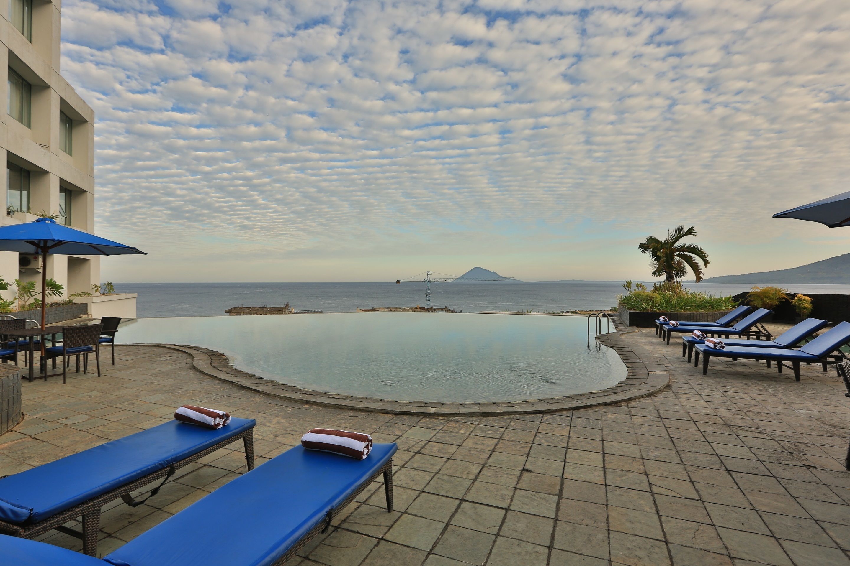 A poolside area with lounge chairs, a pool, and a view of the ocean and a mountain.