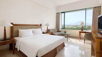 A hotel room with a large bed, two bedside tables with lamps, a small round table, a sofa, a TV, and a large window with a view of the city and mountains.