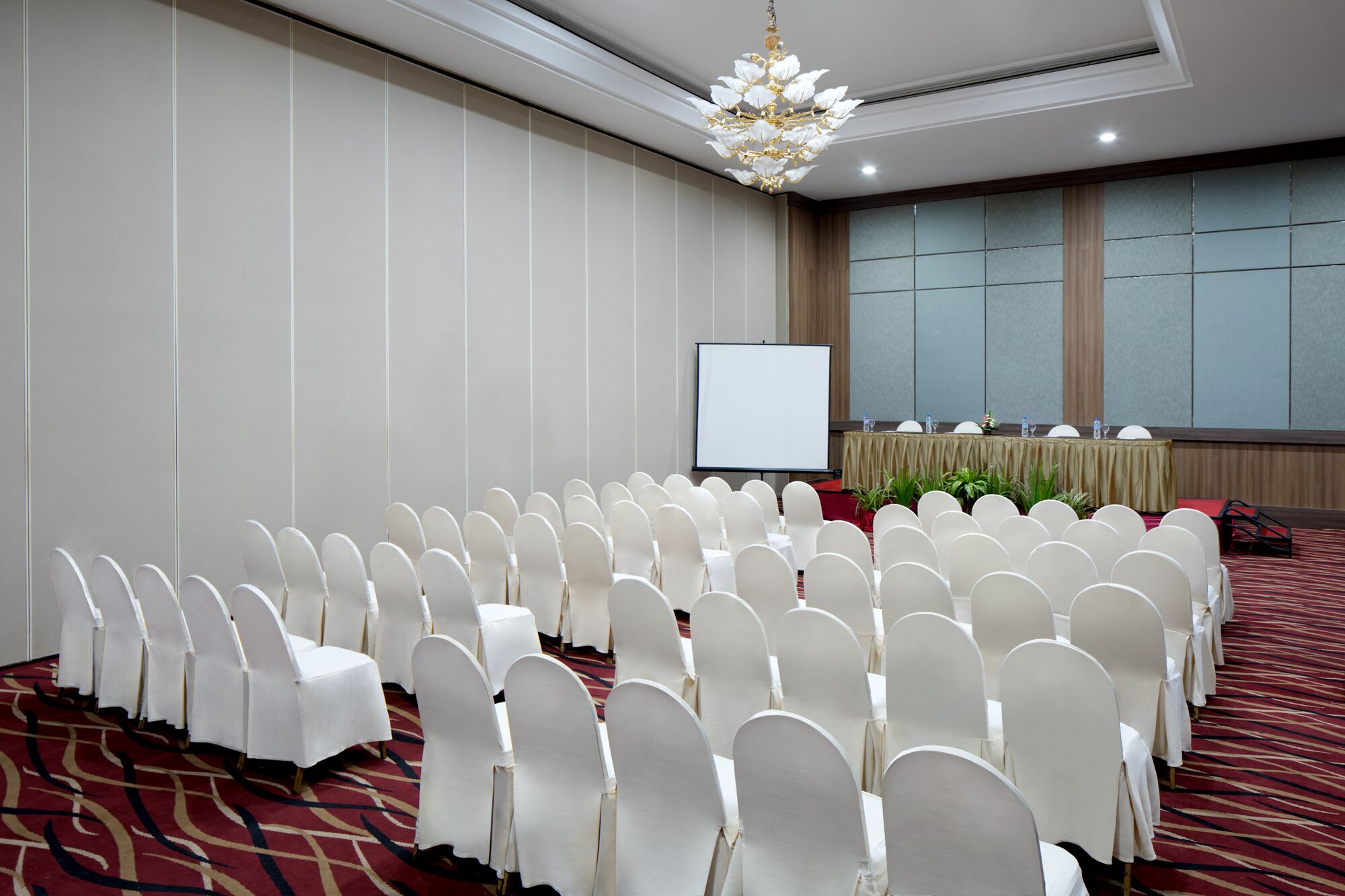 A conference room with rows of white chairs, a projector screen, a table with a tablecloth and water bottles, and a chandelier.