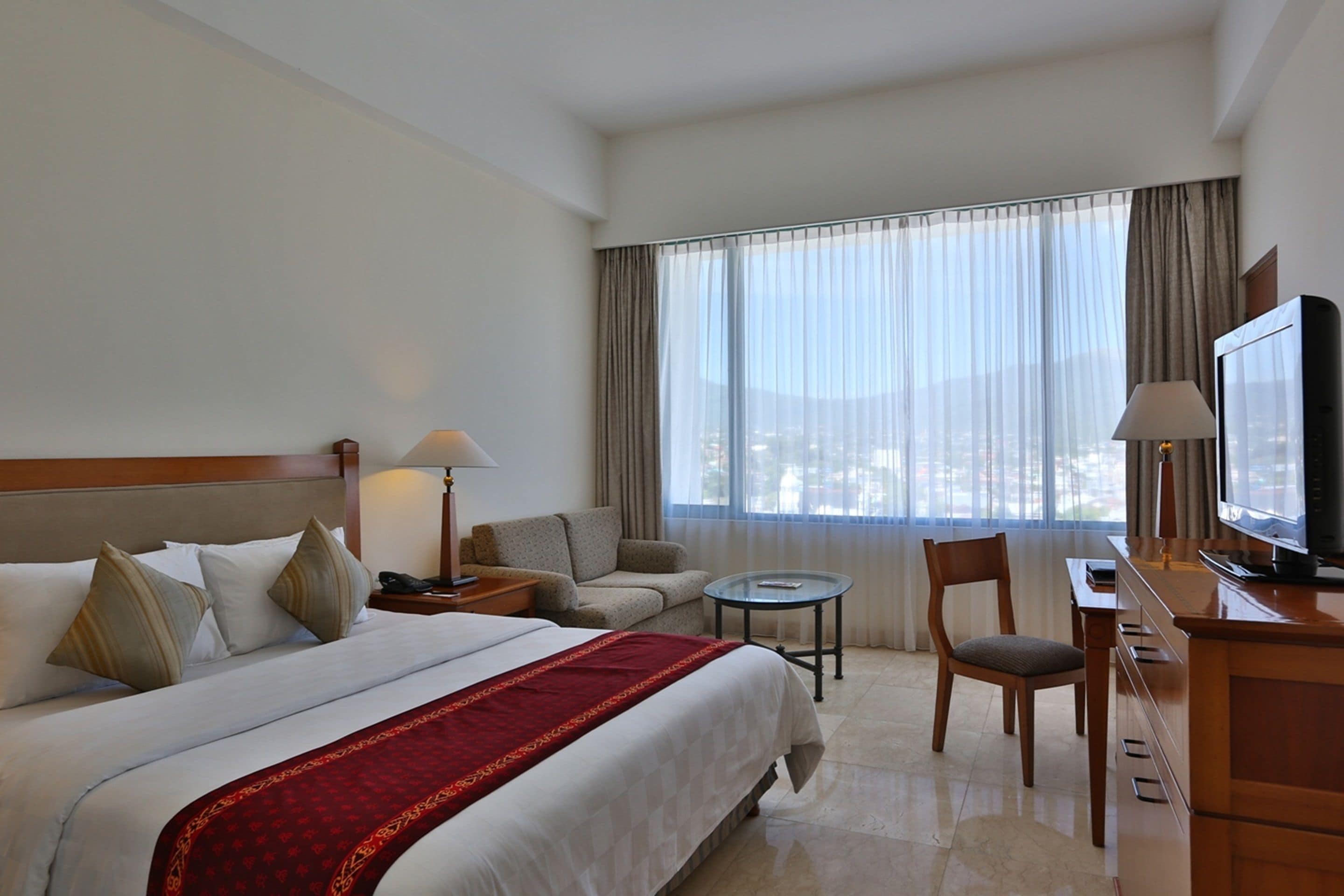 Una habitación de hotel con una cama grande, un sofá, una mesita, un televisor y una ventana con cortinas.