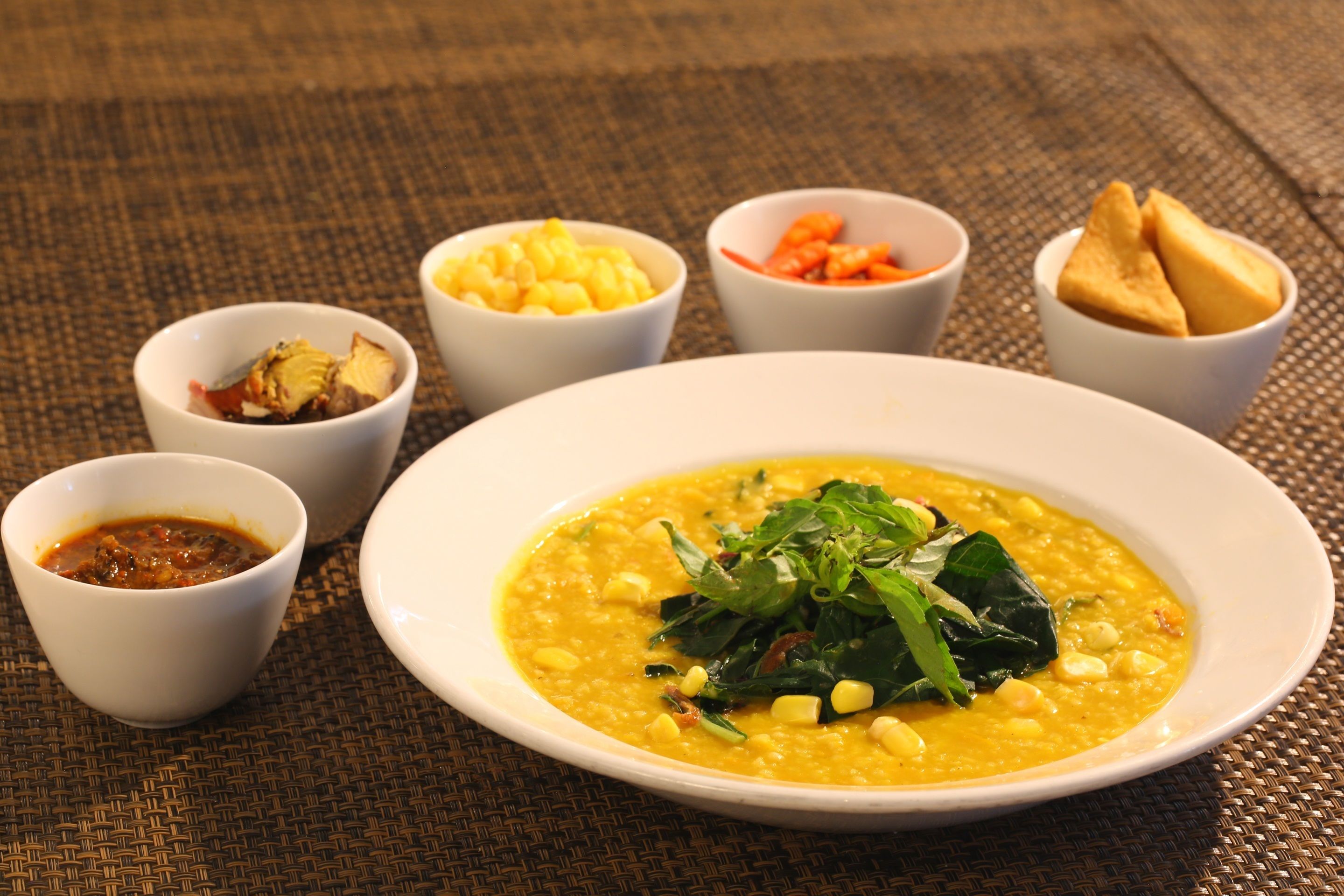 A bowl of lentil soup with corn and greens, accompanied by small bowls of condiments and bread.