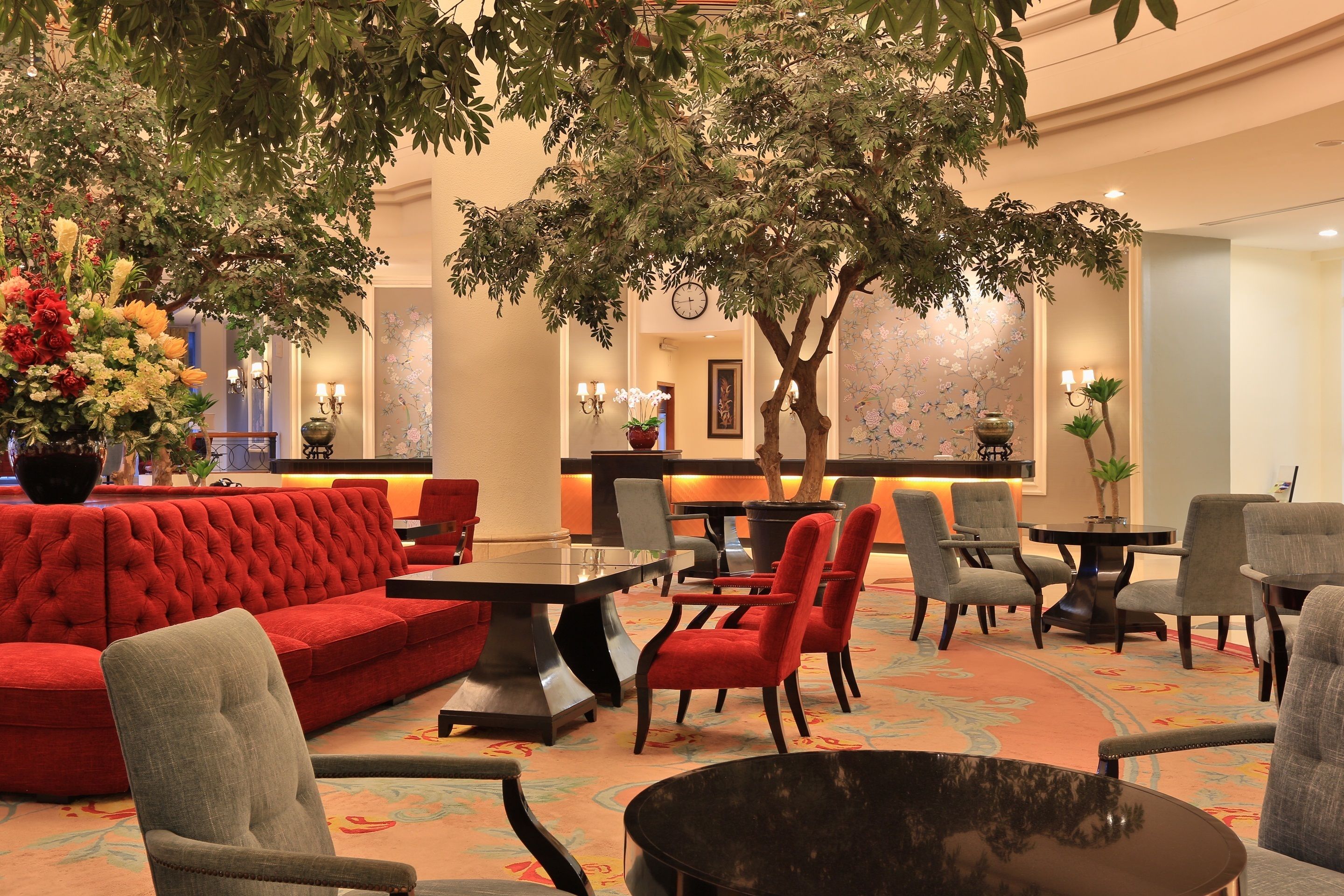 Eine Lobby mit einem roten Sofa, grauen Sesseln, einem runden Tisch und einem großen Baum.