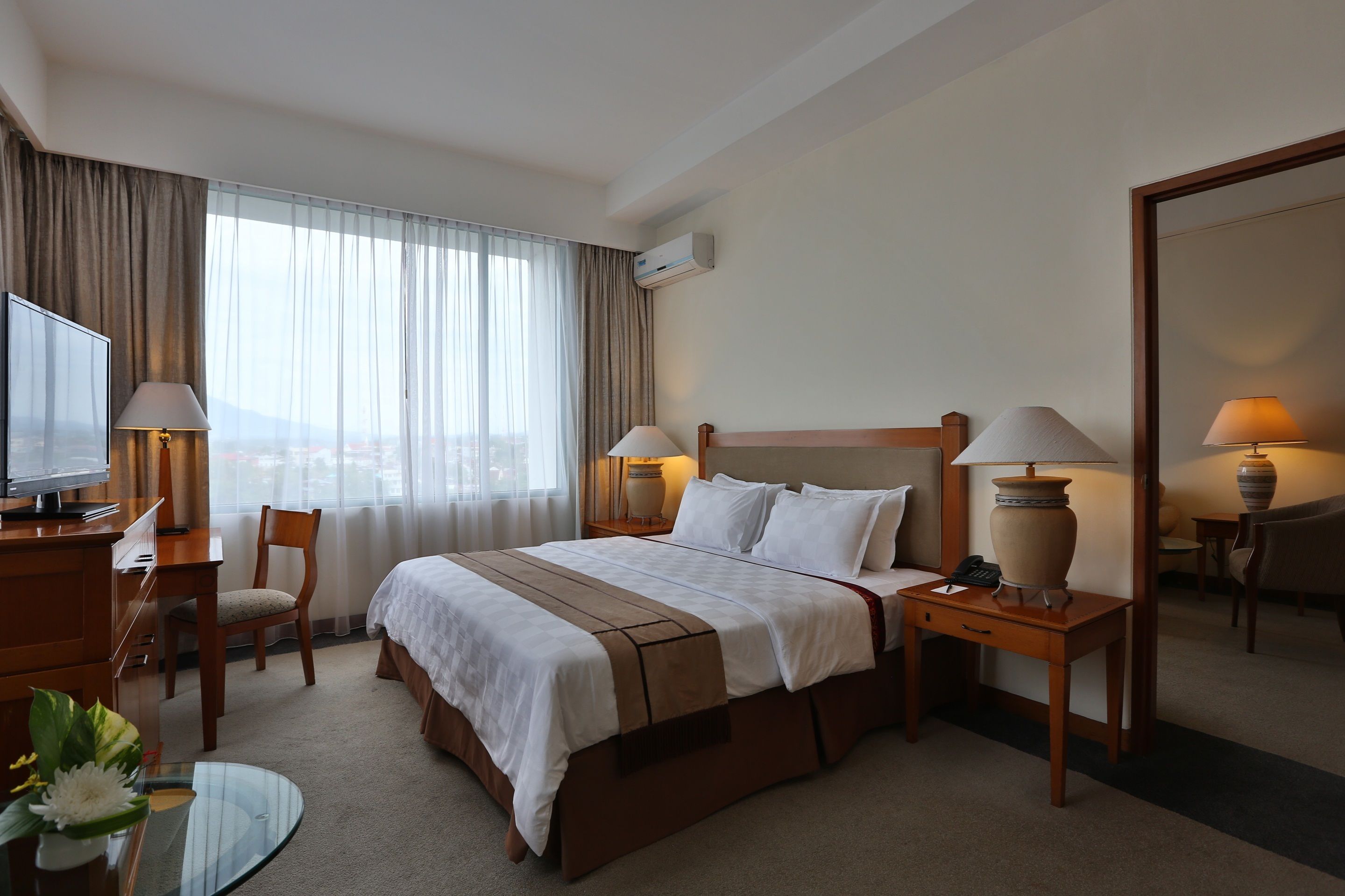 A hotel room with a bed, bedside tables, a desk, a chair, a TV, and a large window with curtains.