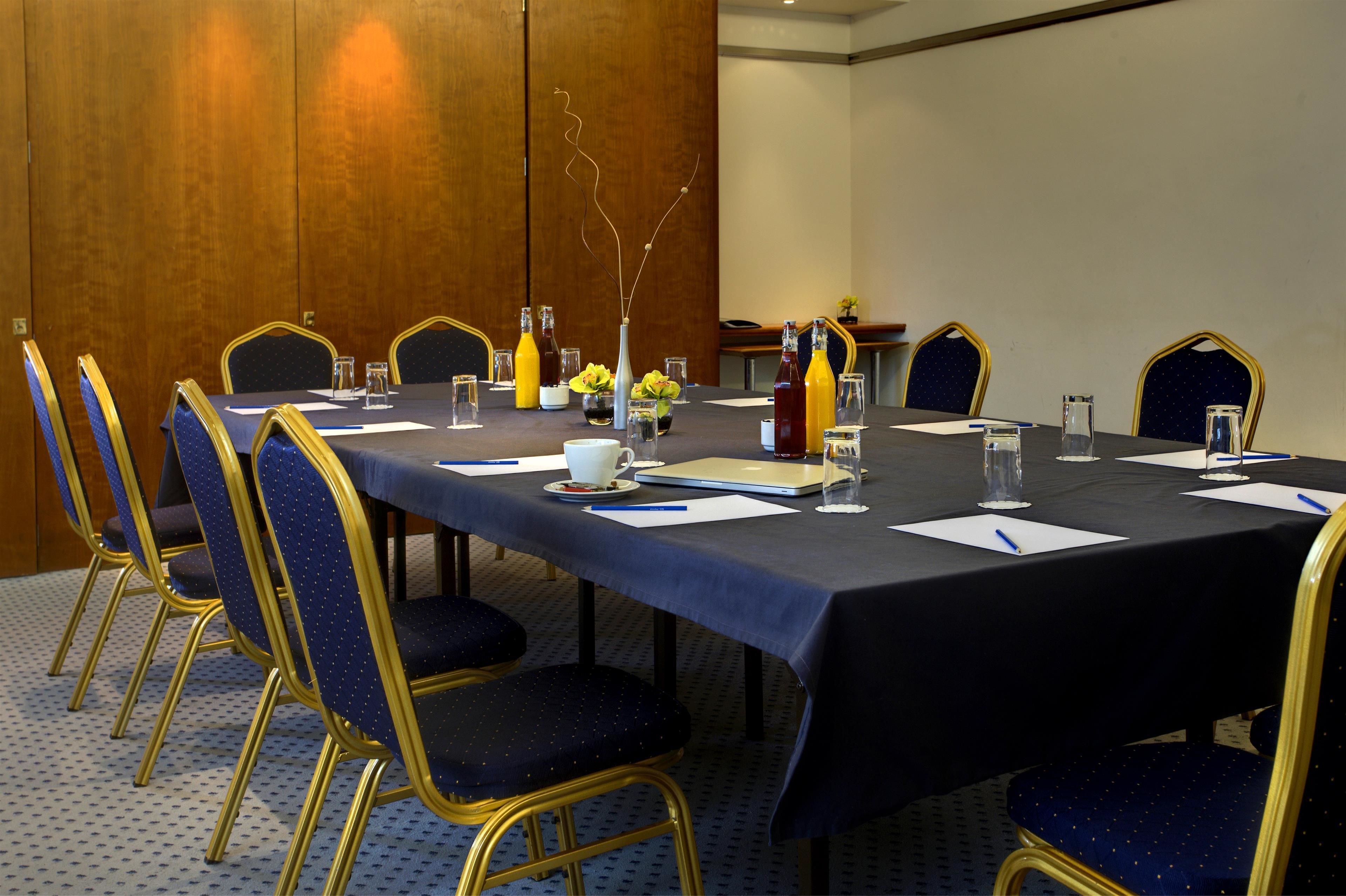 Un salón de conferencias con una mesa larga, sillas y varios objetos como botellas de agua, papeles y un jarrón con flores.