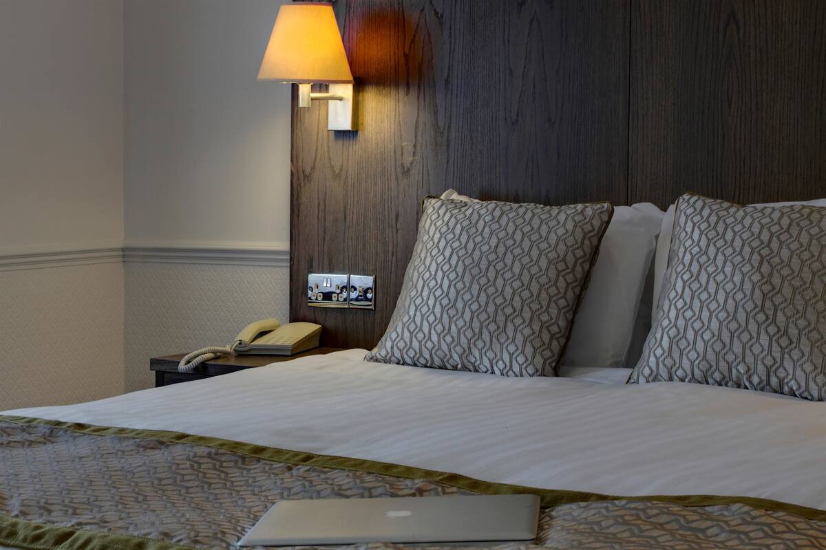 A hotel room with a bed, a bedside lamp, a telephone, and a laptop on the bed.