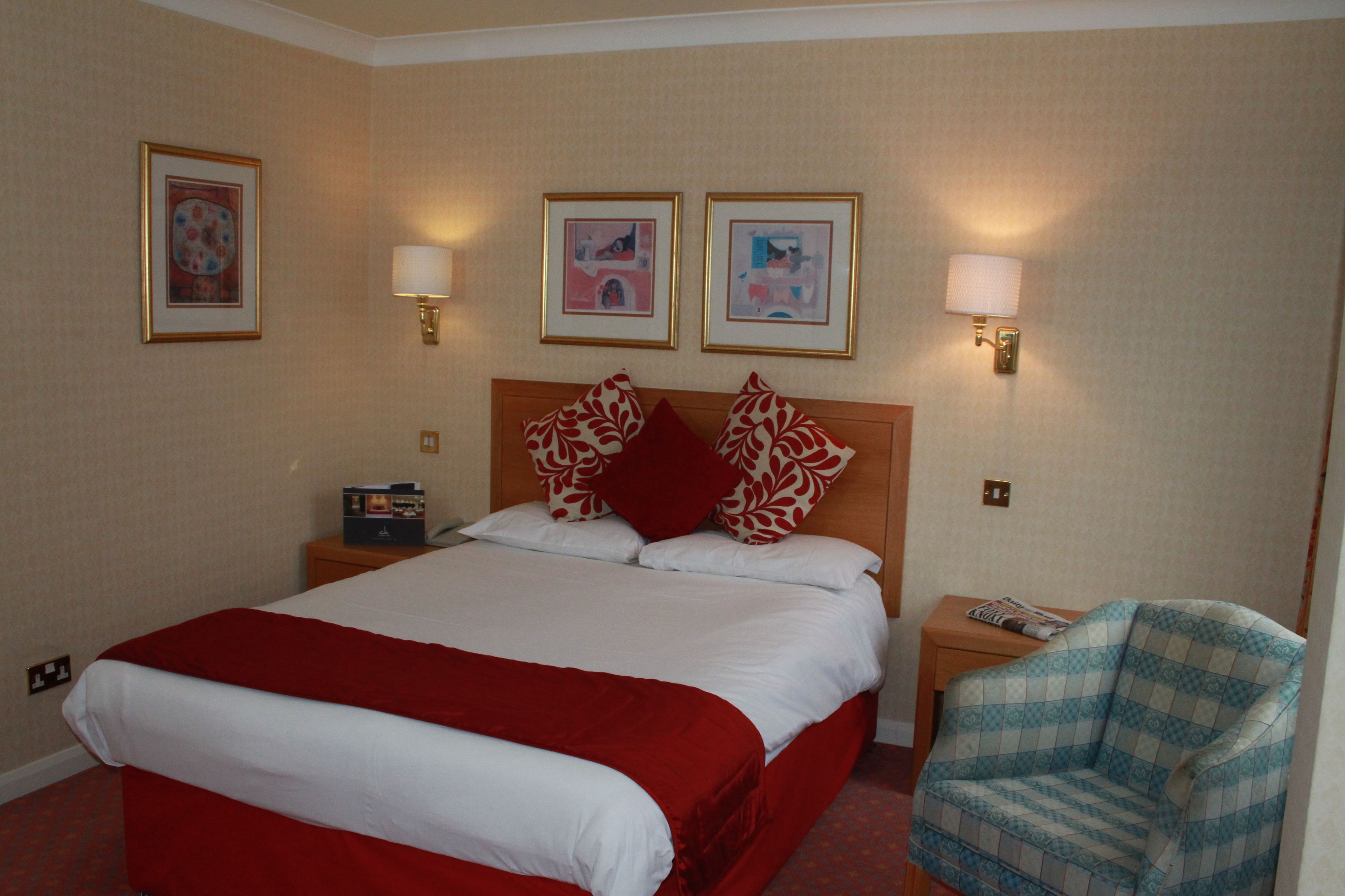 A hotel room with a large bed, two wall-mounted lamps, a chair, and framed pictures on the wall.