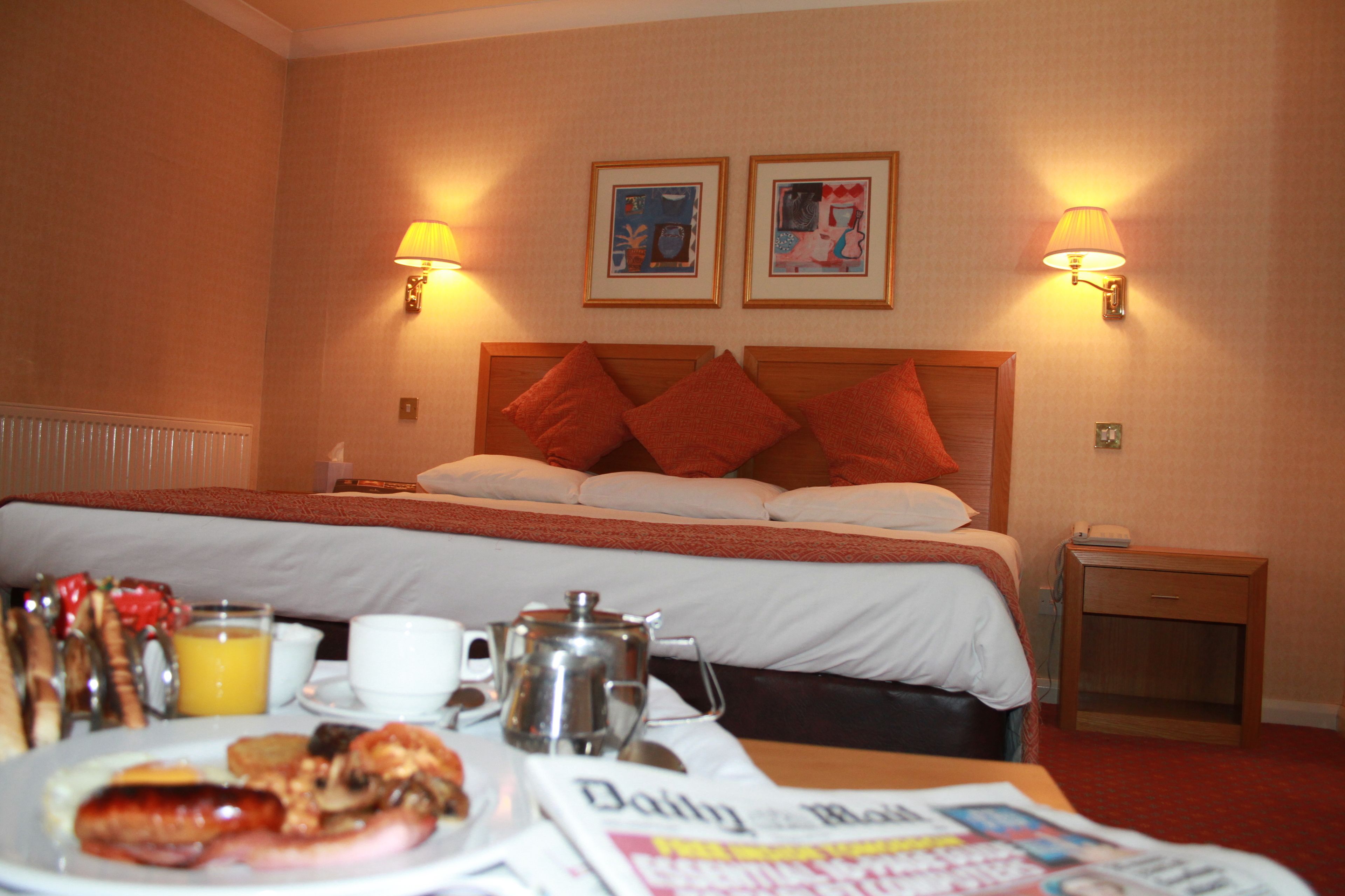A hotel room with a bed, bedside lamps, a nightstand, a table with breakfast items, and two framed pictures on the wall.