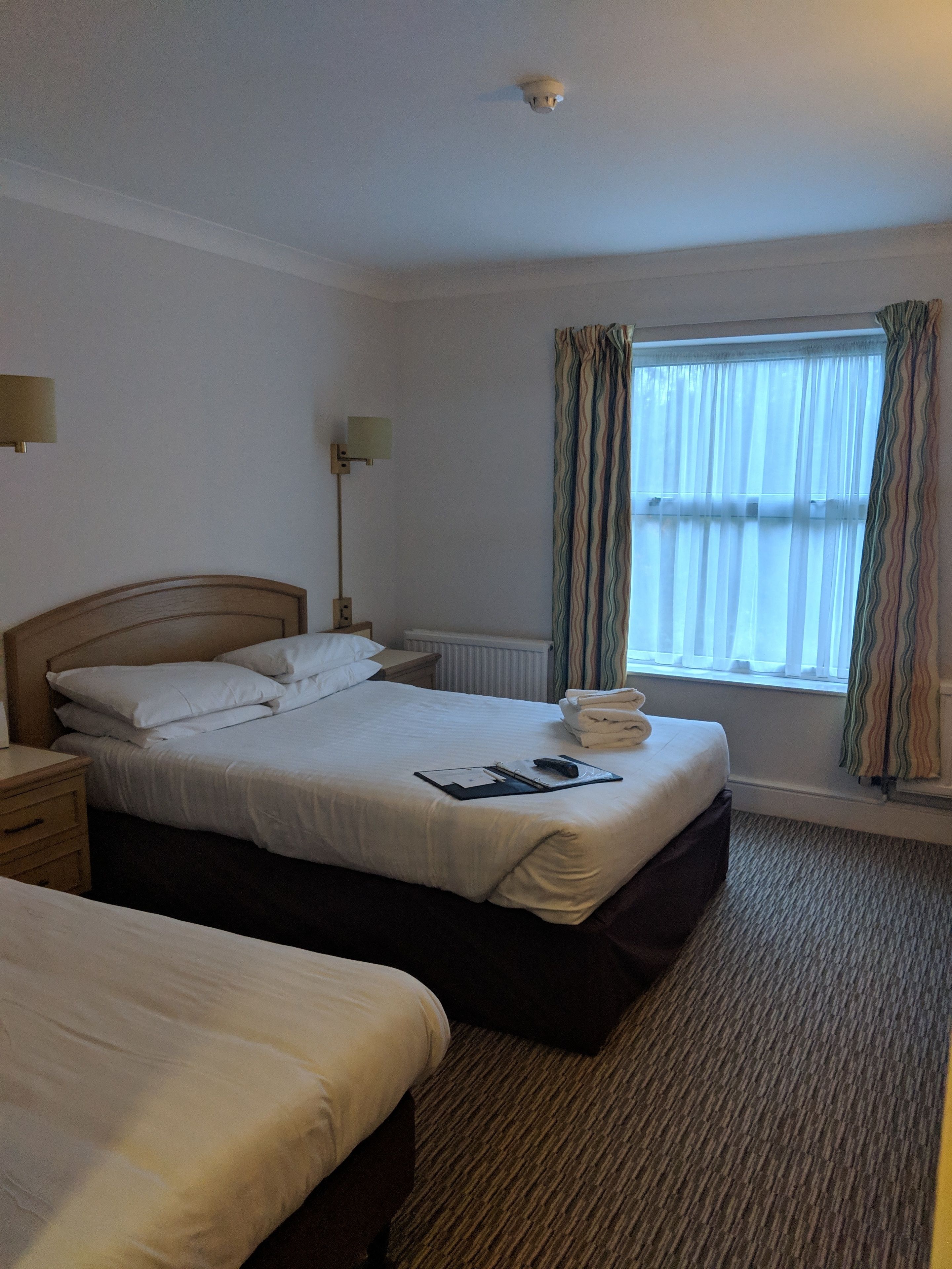 A hotel room with two beds, a bedside lamp, a window with curtains, and a radiator.