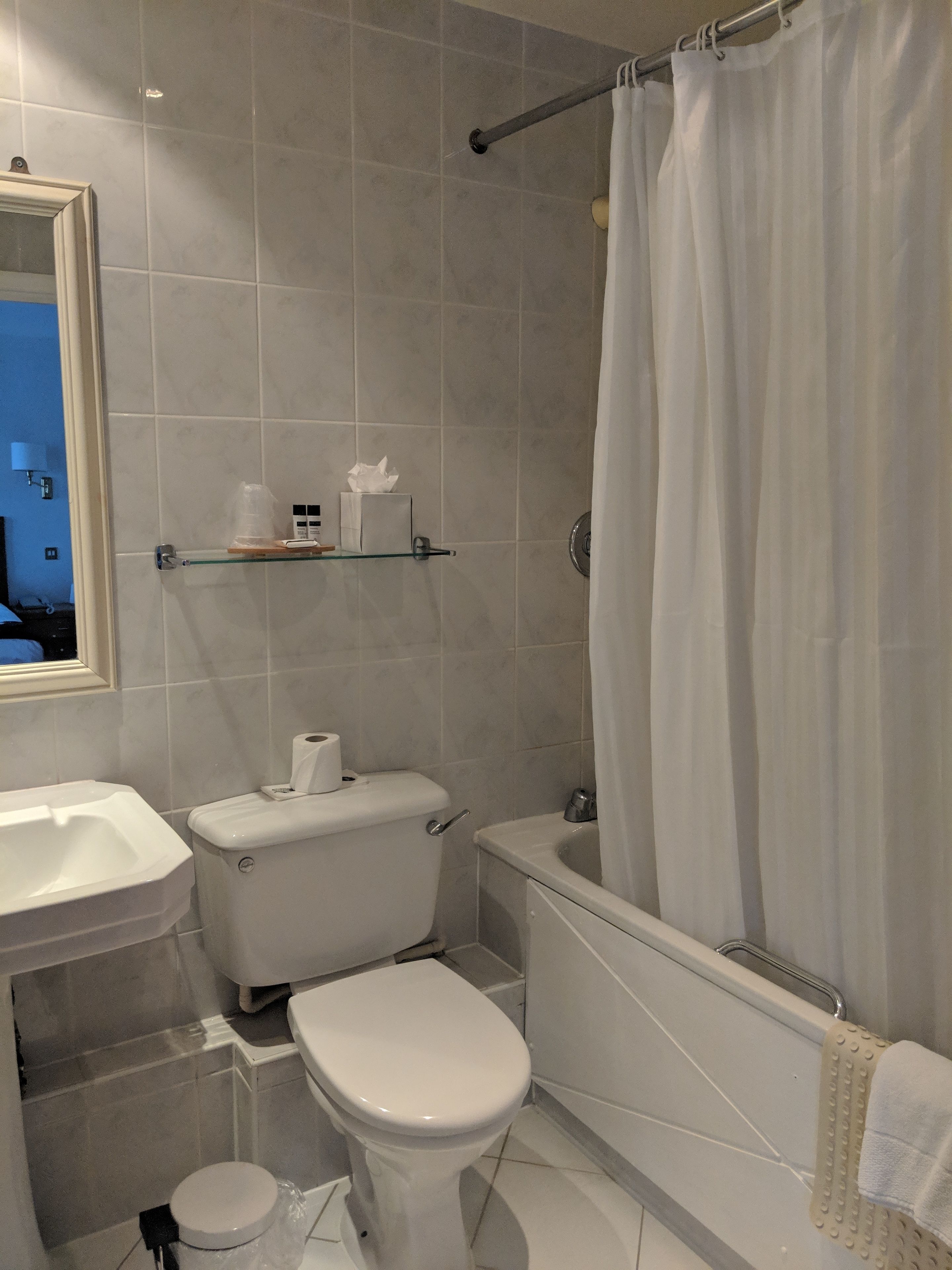 A bathroom with a toilet, sink, and bathtub. There is a towel rack and a glass shelf with toiletries.