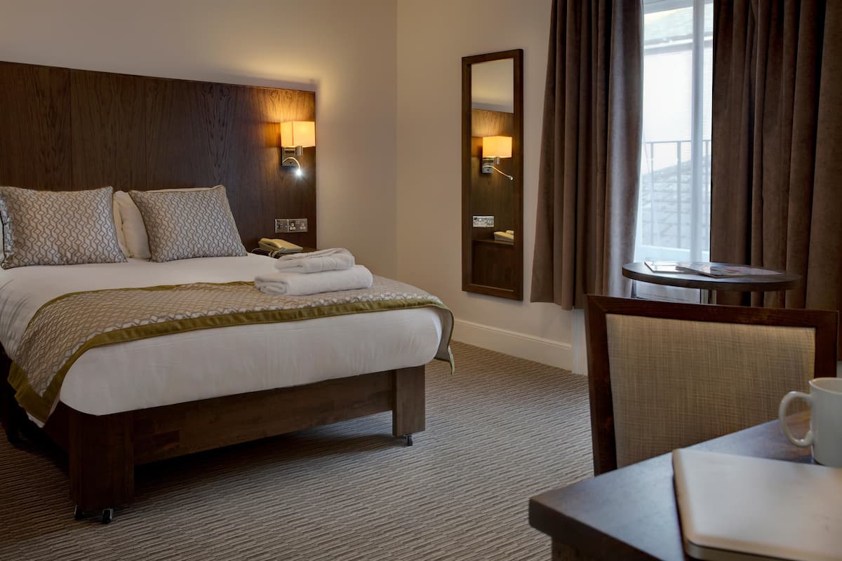 A hotel room with a large bed, two bedside lamps, a mirror, a window with curtains, a small table with a cup, and a chair.