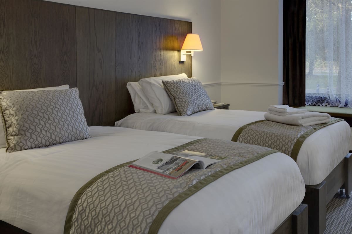 A hotel room with two beds, each with patterned pillows and a magazine on the bed. There is a nightstand with a lamp between the beds.