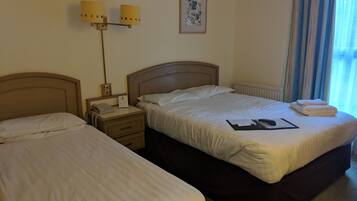 Habitación de hotel con dos camas, una mesita de noche con lámpara, un cuadro enmarcado en la pared y una ventana con cortinas.