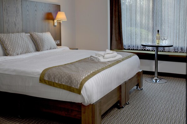 A neatly made bed with a white and gold bedspread, a bedside lamp, and a bottle of wine on a small table beside the window.