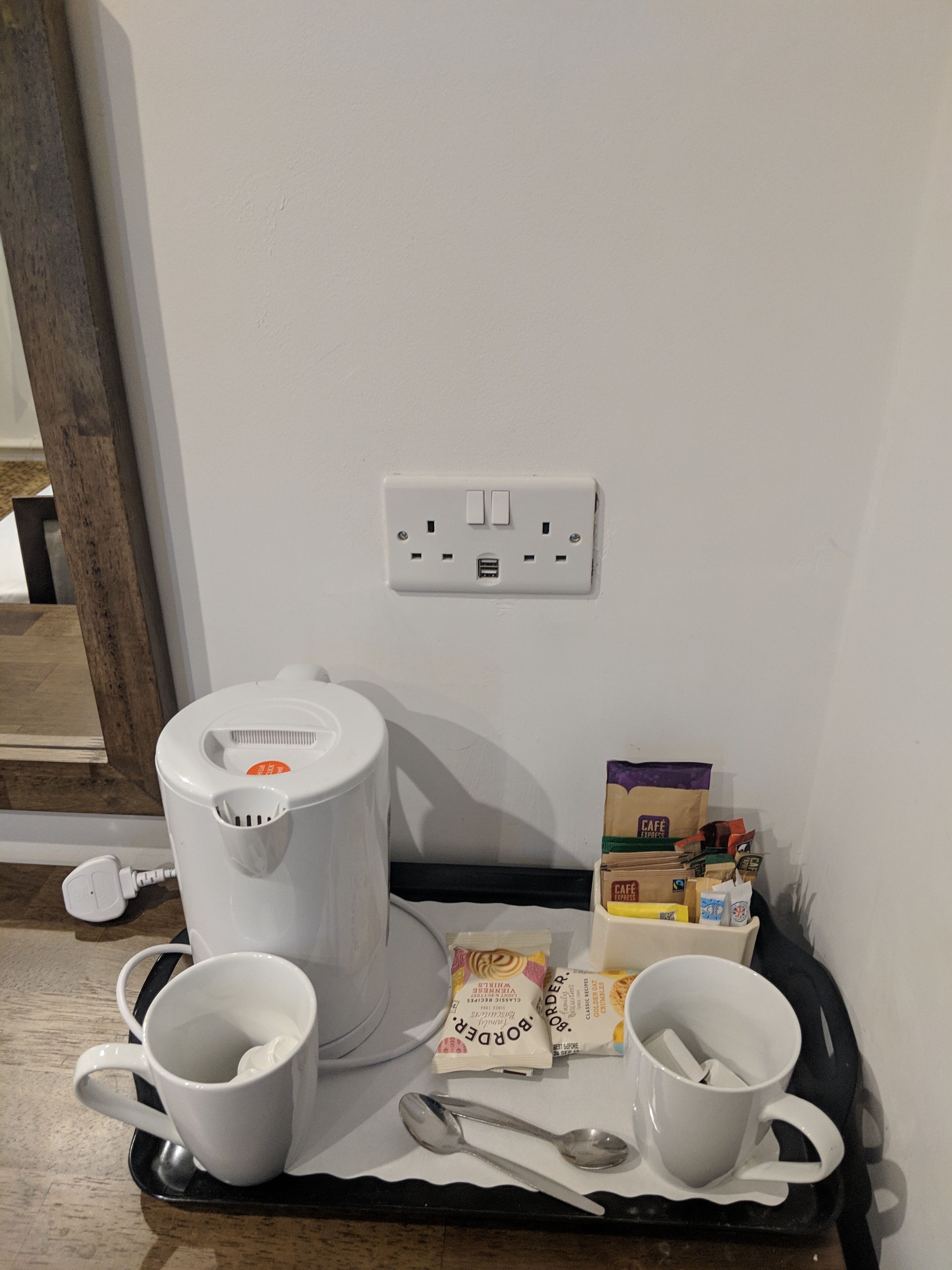 A kitchenette area with a coffee maker, two white mugs, a spoon, and coffee pods on a tray.