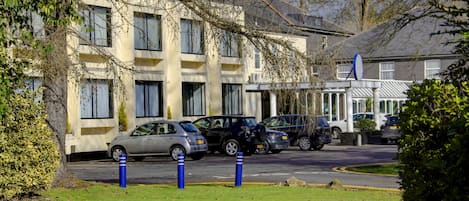 Un aparcamiento con varios coches, un edificio con muchas ventanas y algo de vegetación.