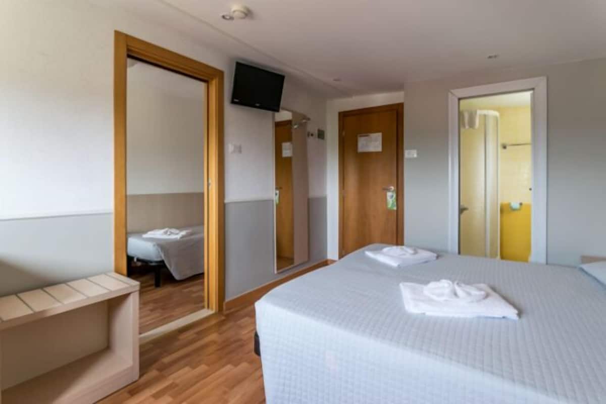 A hotel room with a bed, a TV mounted on the wall, a wooden bench, and a mirror.