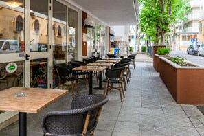 An outdoor cafe with wicker furniture and glass tables, a stone wall, and a view of the street. - Hotel Stresa (Rimini)