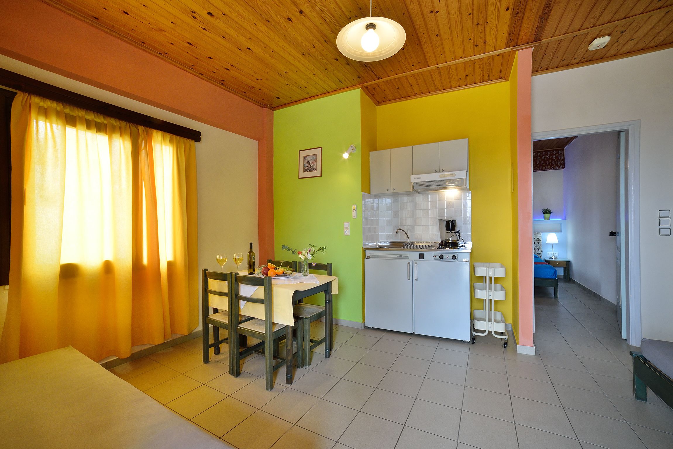 A dining area with a table set for two, a kitchen with white cabinets, and a hallway leading to another room.