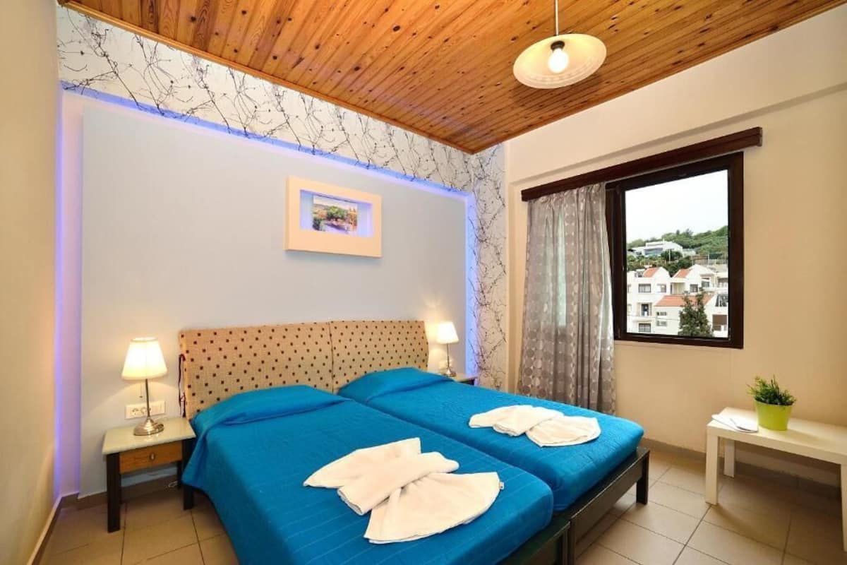 Une chambre d’hôtel avec deux lits, un plafond en bois, une fenêtre donnant sur des immeubles et une petite table avec une plante.