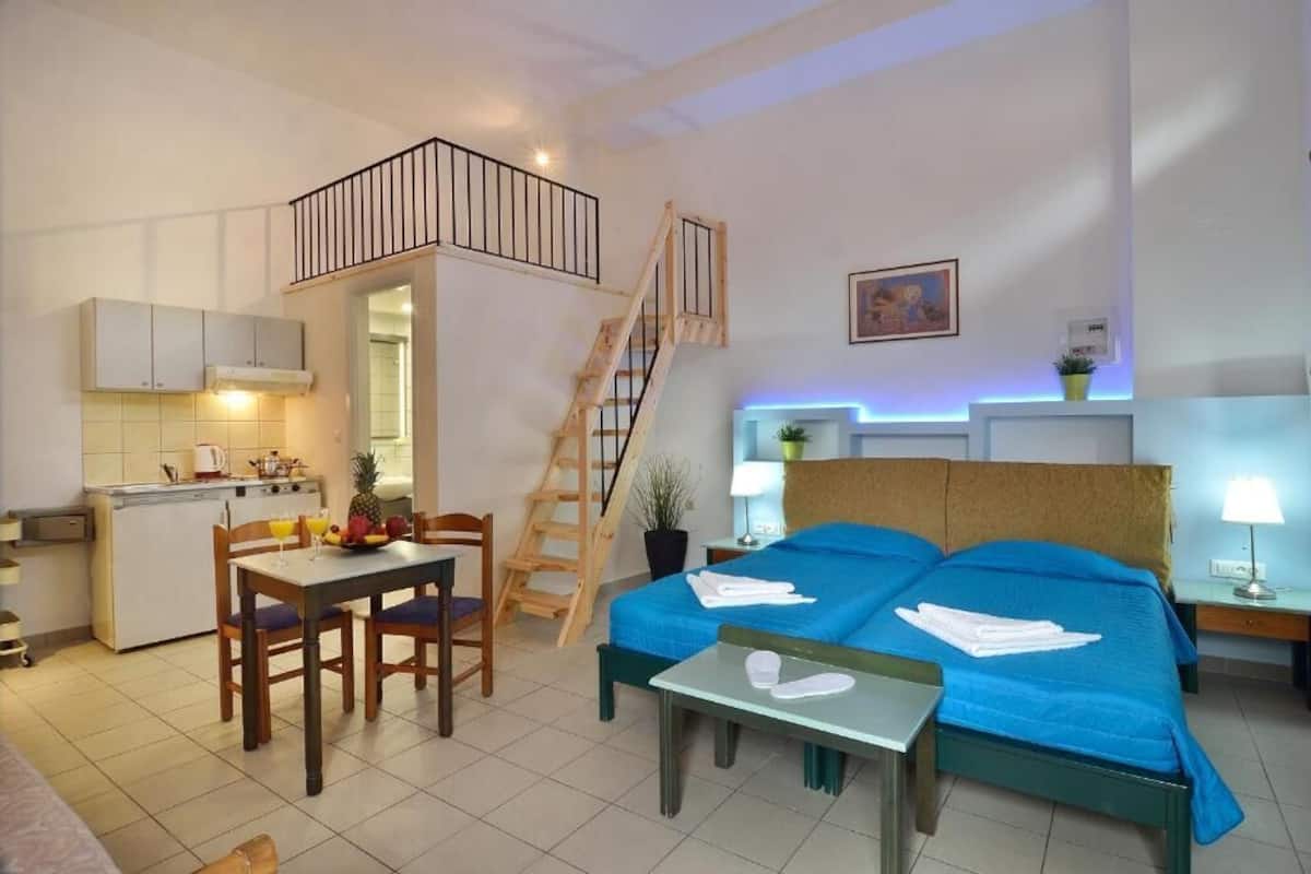 Une chambre d’hôtel compacte comprenant un coin cuisine, un espace repas, un lit avec une literie bleue et un escalier.