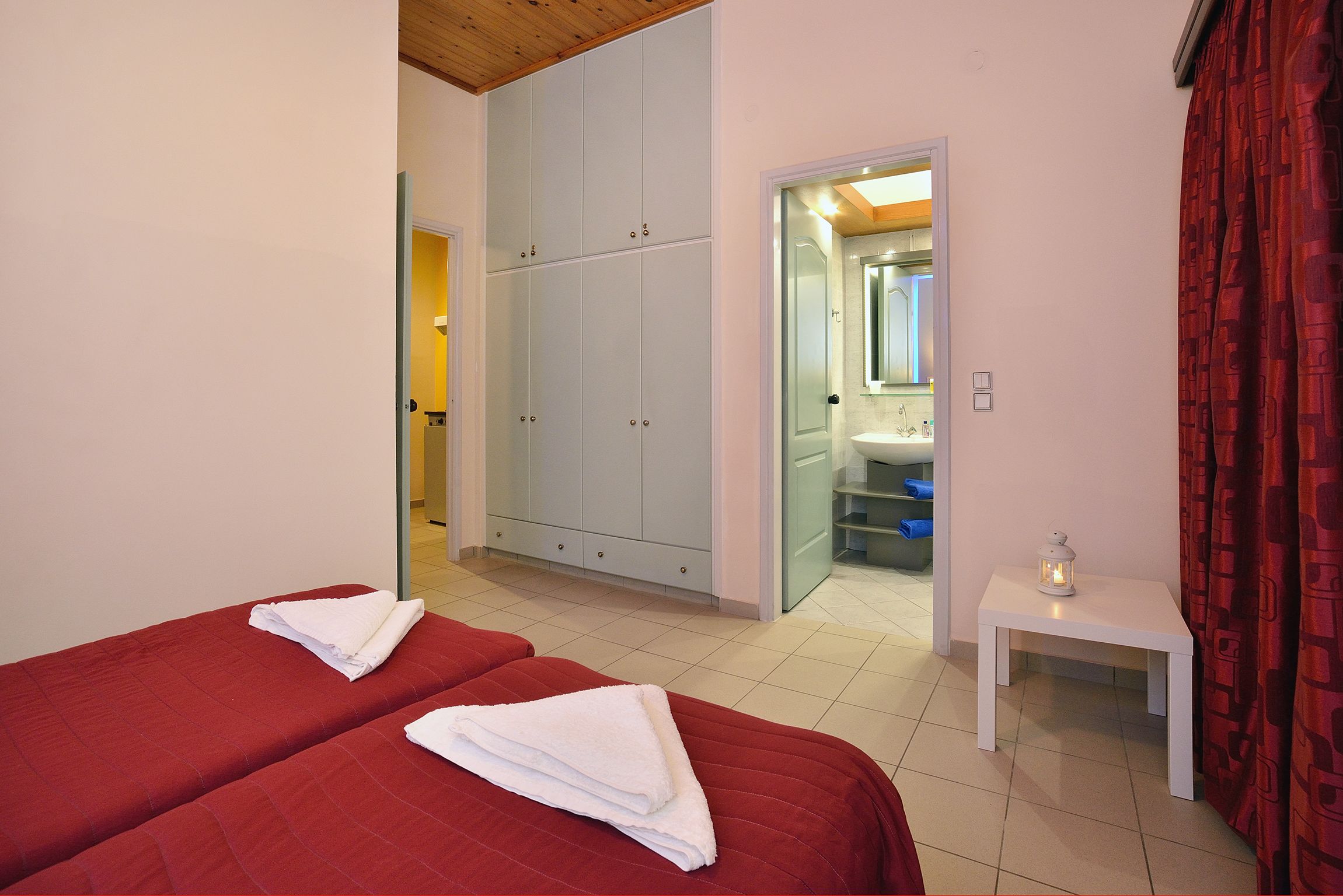 A hotel room with two beds, a wardrobe, a bathroom with a sink, and a small table with a lantern.