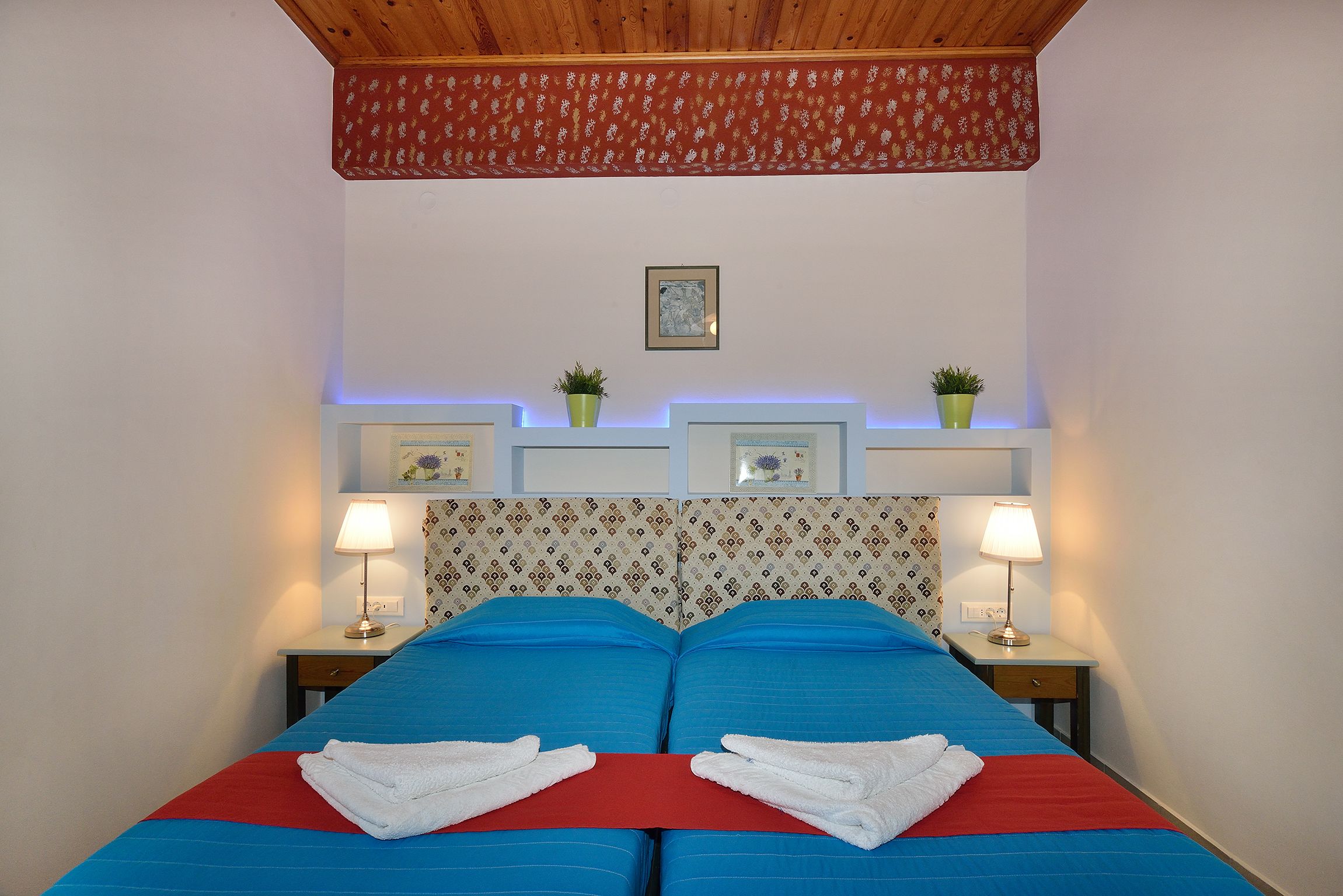 A double bed with blue and red bedding, two bedside tables with lamps, a floral-patterned headboard, and a small framed picture on the wall.
