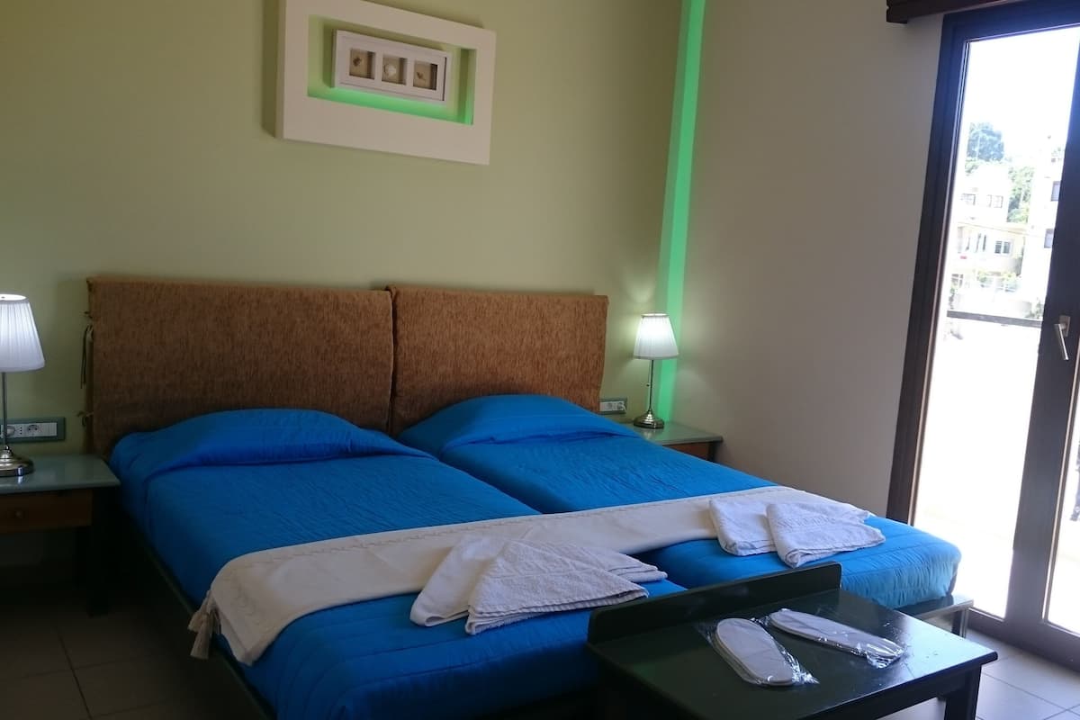 Une chambre d’hôtel avec un lit double, deux lampes de chevet, une petite table avec un téléphone et une serviette, et une vue sur les bâtiments par la fenêtre.