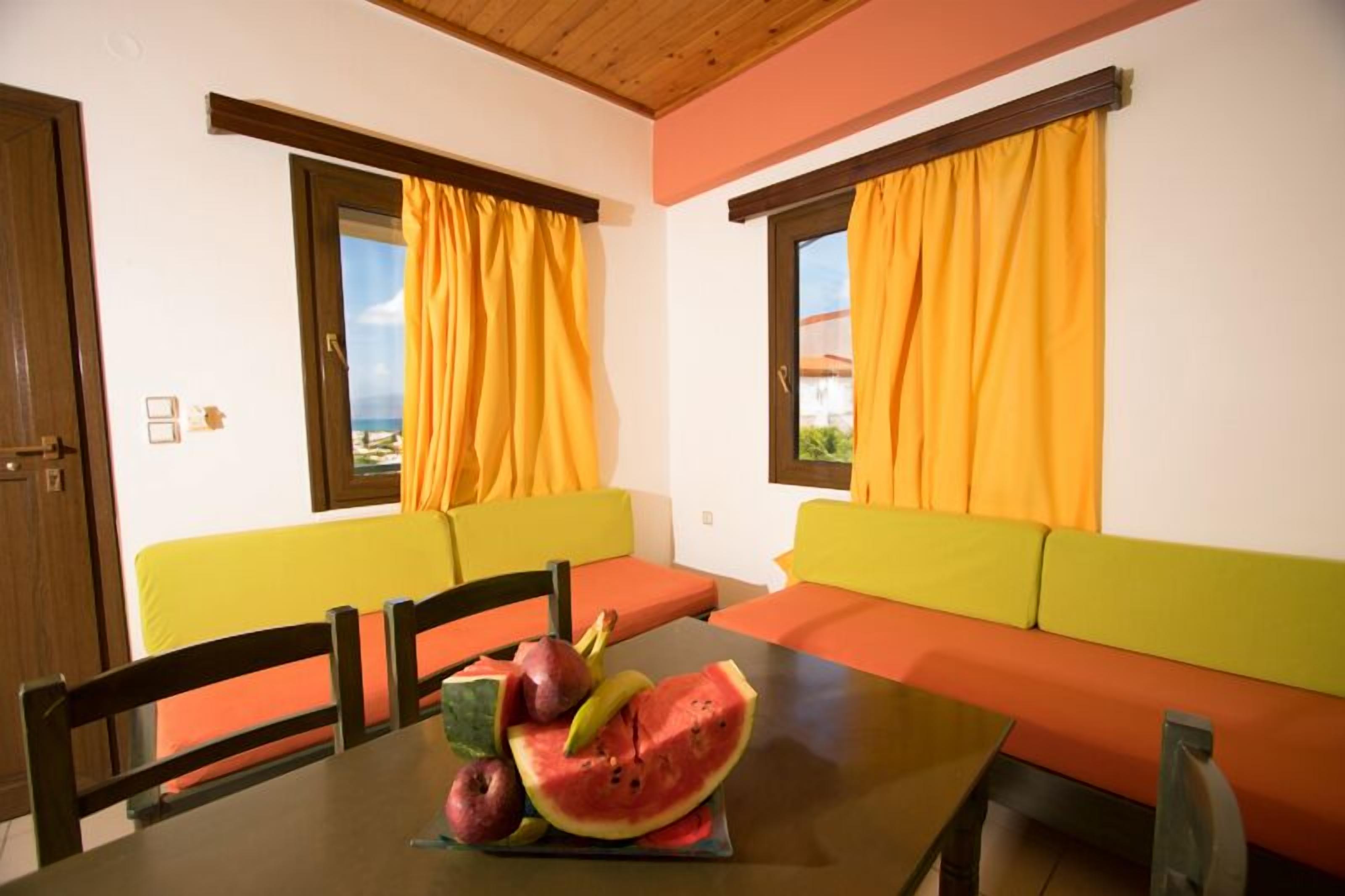 A dining area with a table set for two, a fruit platter, and two sofas with orange cushions.