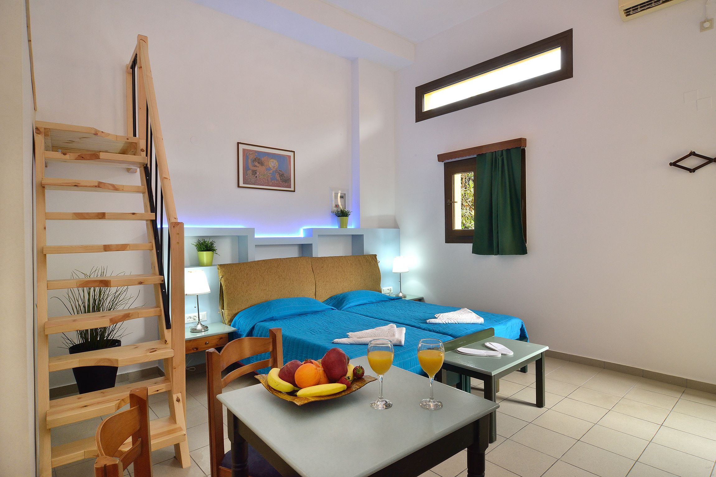 A hotel room with a bed, a desk with a fruit bowl and glasses, a chair, a ladder, a window with curtains, and a wall-mounted air conditioner.