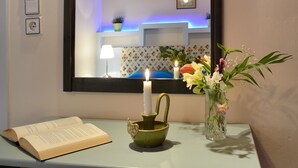 Una stanza con un comodino, una candela accesa, un libro aperto, un vaso con fiori e uno specchio che riflette una mensola illuminata di blu e un letto.