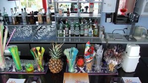 A bar area with various bottles and glasses on shelves, a coffee machine, and promotional materials on the counter. - Memories Apartments (Hersonissos)