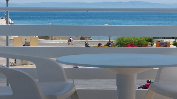 Un balcón con sillas y una mesa blancos, con vistas a la costa y al mar, con montañas lejanas.