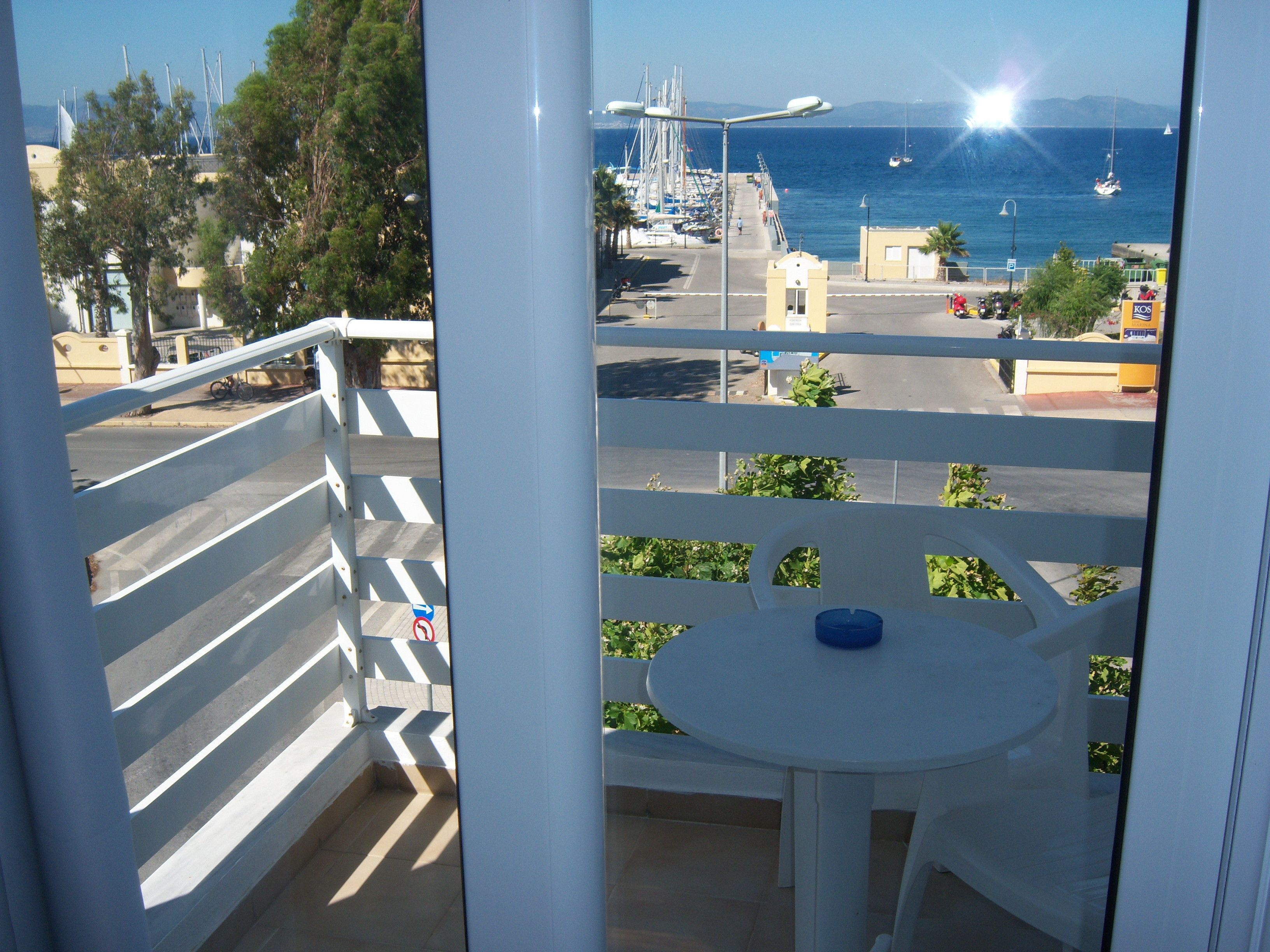 A balcony with a white railing, a small table with a blue object, and a view of a marina with boats and a clear sky.