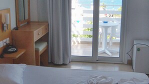 Una habitación de hotel con cama, escritorio, espejo, ventana con vista al mar y barcos, y balcón con sillas y mesa blancos.