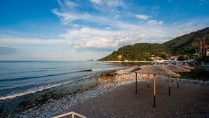 A beach with pebbles, a clear sky, and a few umbrellas. - Kamelia Hotel (SARONIC ISLANDS)