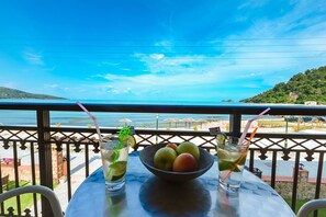A table with a bowl of fruit and two drinks, overlooking a beach with umbrellas and a clear sky. - Kamelia Hotel (SARONIC ISLANDS)