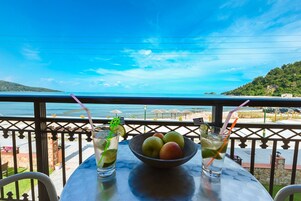 A table with a bowl of fruit and two drinks, overlooking a beach with umbrellas and a clear sky.