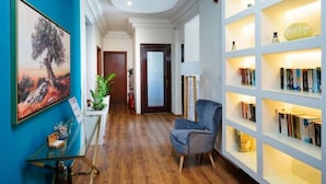 A hallway with a blue wall, a glass table with a plant, a blue armchair, and a bookshelf with books. - Kamelia Hotel (SARONIC ISLANDS)