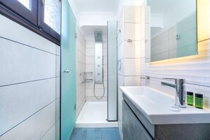 A modern bathroom with a glass-enclosed shower, a white sink with a chrome faucet, and a large mirror above the sink. - Kamelia Hotel (SARONIC ISLANDS)